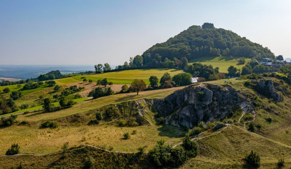Grüne Hügel und Bäume unter klarem Himmel, mit einem markanten Felsen im Vordergrund. Im Hintergrund ist ein bewaldeter Hügel zu sehen., © TMBW Grüne Hügel und Bäume unter klarem Himmel, mit einem markanten Felsen im Vordergrund. Im Hintergrund ist ein bewaldeter Hügel zu sehen., © TMBW