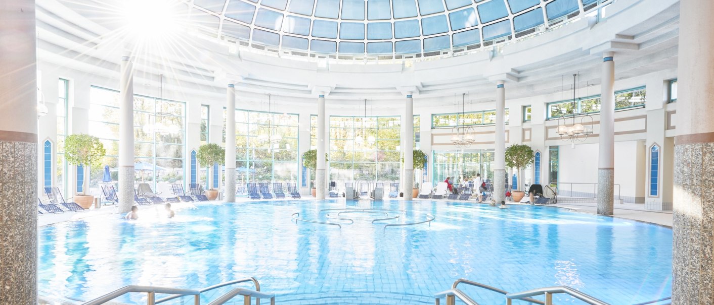 Großes Innenbecken mit Glaskuppel, Sonnenlicht strahlt herein. Menschen schwimmen, Liegestühle und Pflanzen am Rand., © Mineraltherme Böblingen