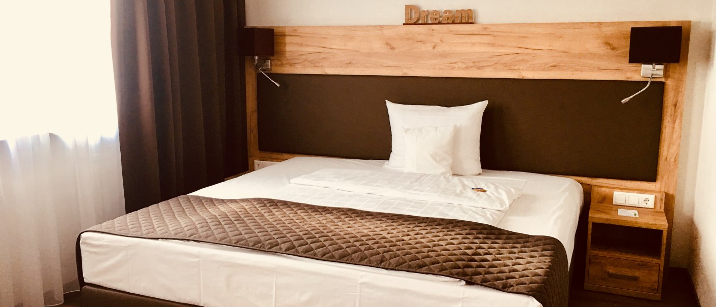 Modernes Hotelzimmer mit Doppelbett, braunen Vorhängen und Holzmöbeln. Auf dem Kopfteil steht 'Dream'. Helle, gemütliche Atmosphäre., © TOMAS Modernes Hotelzimmer mit Doppelbett, braunen Vorhängen und Holzmöbeln. Auf dem Kopfteil steht 'Dream'. Helle, gemütliche Atmosphäre., © TOMAS