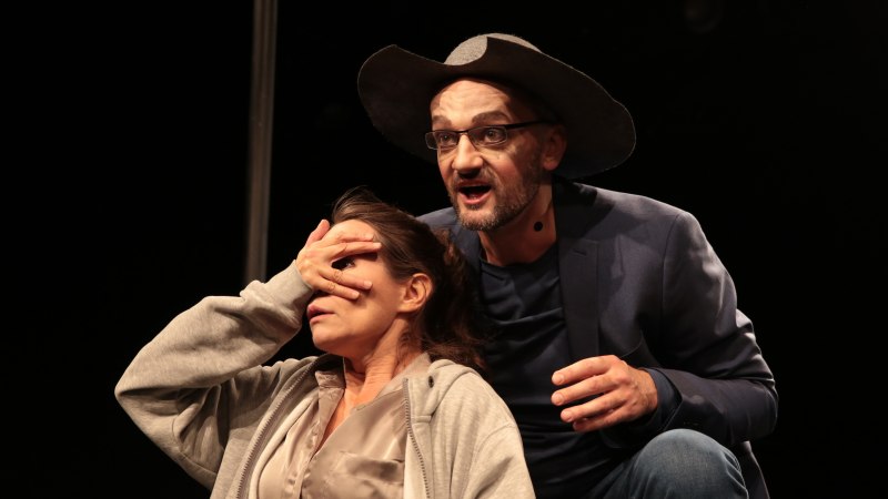 Ein Mann mit Hut und Brille spricht auf der Bühne, während er die Augen einer Frau verdeckt. Beide tragen Mikrofone., © Theater tri-bühne, Stefan Kirchknopf Ein Mann mit Hut und Brille spricht auf der Bühne, während er die Augen einer Frau verdeckt. Beide tragen Mikrofone., © Theater tri-bühne, Stefan Kirchknopf
