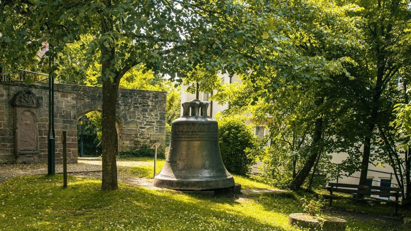 Eine gro&szlig;e Glocke steht im Garten der Stiftskirche Herrenberg, umgeben von B&auml;umen und einer alten Steinmauer. Sonnenlicht f&auml;llt durch das Bl&auml;tterdach., &copy; Stuttgart-Marketing GmbH, Sarah Schmid