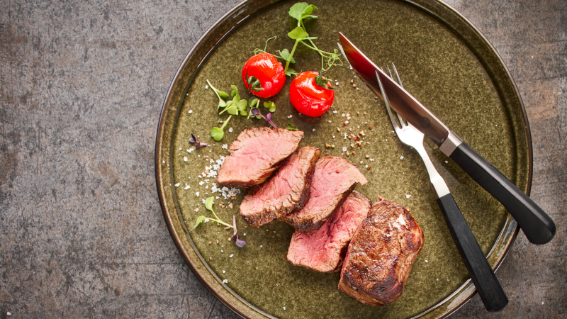 Ein Teller mit geschnittenem Steak, zwei Kirschtomaten und Besteck auf einem rustikalen Untergrund., &copy; MAREDO