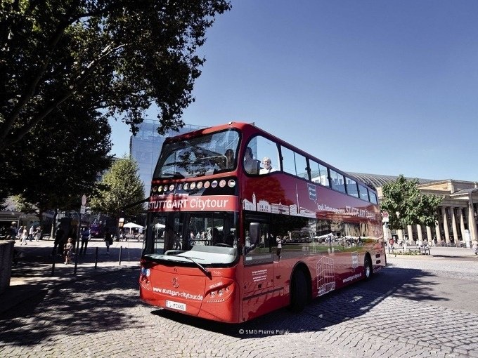 Roter Doppeldeckerbus der Stuttgart Citytour auf gepflasterter Straße, umgeben von Bäumen und Gebäuden., © SMG, Pierre Polak Roter Doppeldeckerbus der Stuttgart Citytour auf gepflasterter Straße, umgeben von Bäumen und Gebäuden., © SMG, Pierre Polak