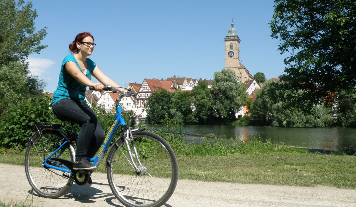 Eine Frau fährt Fahrrad am Neckar in Nürtingen. Im Hintergrund sind eine Kirche und Fachwerkhäuser zu sehen., © Stadt Nürtingen Eine Frau fährt Fahrrad am Neckar in Nürtingen. Im Hintergrund sind eine Kirche und Fachwerkhäuser zu sehen., © Stadt Nürtingen