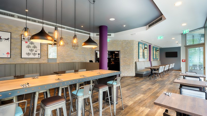 Moderner Frühstücksbereich mit langen Holztischen, Metallstühlen und stilvollen Hängelampen. Steinwände und große Fenster schaffen eine einladende Atmosphäre., © Premier Inn Germany Moderner Frühstücksbereich mit langen Holztischen, Metallstühlen und stilvollen Hängelampen. Steinwände und große Fenster schaffen eine einladende Atmosphäre., © Premier Inn Germany