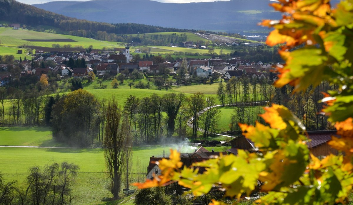 Grünes Tal mit Dorf im Hintergrund, umgeben von Hügeln. Im Vordergrund bunte Herbstblätter. Rauch steigt aus einem Schornstein auf., © Landkreis Göppingen
