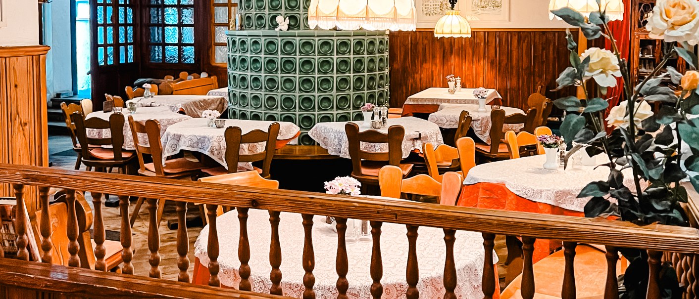 Gemütlicher Gastraum mit Holzmöbeln, einem grünen Kachelofen und dekorativen Tischdecken. Hängelampen und Blumenarrangements ergänzen das Ambiente., © Weinstube Kachelofen