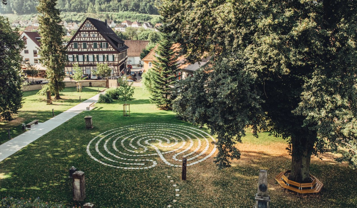 Ein Labyrinth aus Steinen im Kirchgarten, umgeben von Bäumen und einem Fachwerkhaus im Hintergrund. Ein ruhiger, grüner Ort., © Tourismus Ostalb