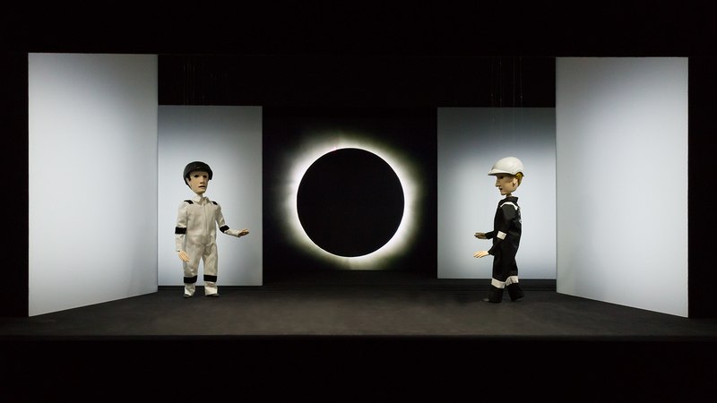 Zwei Puppen in weißen und schwarzen Anzügen stehen auf einer Bühne vor einer Sonnenfinsternis. Die Szene ist minimalistisch gestaltet., © LIMA-Theater Zwei Puppen in weißen und schwarzen Anzügen stehen auf einer Bühne vor einer Sonnenfinsternis. Die Szene ist minimalistisch gestaltet., © LIMA-Theater