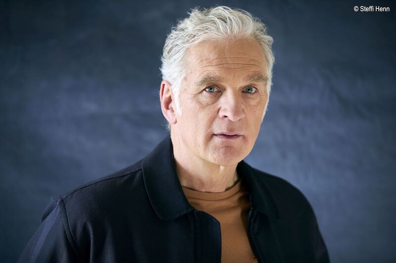 Ein &auml;lterer Mann mit grauen Haaren und ernstem Blick steht vor einem blauen Hintergrund. Er tr&auml;gt eine dunkle Jacke und ein braunes Shirt., &copy; Stadthalle Leonberg