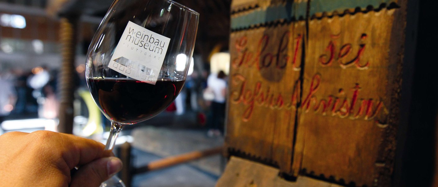 Ein Glas Rotwein mit dem Logo des Weinbaumuseums Stuttgart, im Hintergrund eine alte Holzpresse mit der Inschrift 'Gelobt sei Jesus Christus'., © Stuttgart-Marketing GmbH