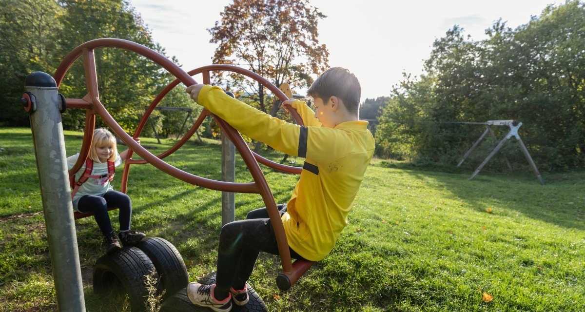 Zwei Kinder spielen auf einem Klettergerüst aus Reifen und Metallstangen auf einem grünen Spielplatz. Im Hintergrund sind Bäume und eine Schaukel zu sehen., © Foto Thomas Zehnder Zwei Kinder spielen auf einem Klettergerüst aus Reifen und Metallstangen auf einem grünen Spielplatz. Im Hintergrund sind Bäume und eine Schaukel zu sehen., © Foto Thomas Zehnder