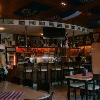 Gemütliche Bar mit Holzmöbeln, Flaggen an der Decke und dekorativen Postern. Die Theke ist gut bestückt, und es gibt mehrere Sitzmöglichkeiten., © cArchie Gemütliche Bar mit Holzmöbeln, Flaggen an der Decke und dekorativen Postern. Die Theke ist gut bestückt, und es gibt mehrere Sitzmöglichkeiten., © cArchie