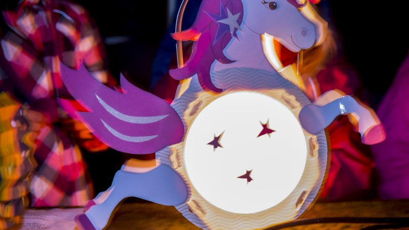 Bunte Einhorn-Laterne mit leuchtendem Körper und Sternen, umgeben von unscharfen Personen in bunter Kleidung. Stimmungsvoll und festlich., © Torsten Wenzler