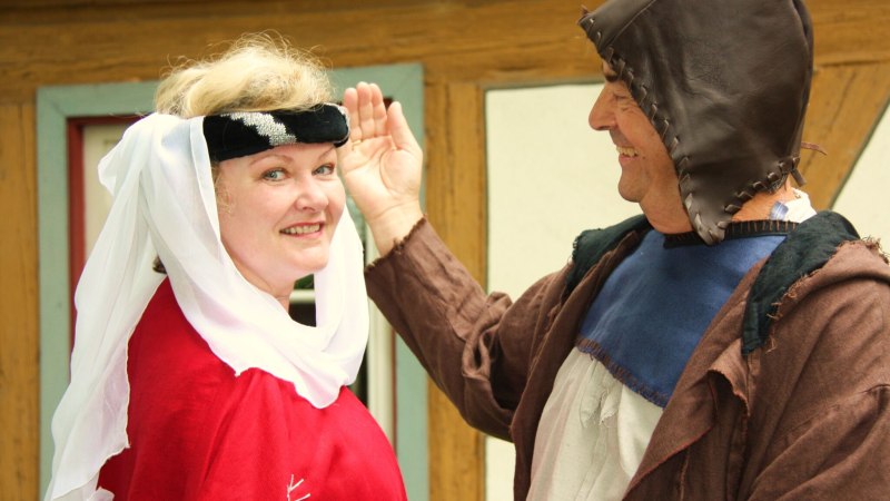 Zwei Personen in mittelalterlicher Kleidung lächeln einander an. Die Frau trägt ein rotes Gewand und eine weiße Kopfbedeckung, der Mann eine braune Kapuze., © Für die Bildrechte: Amt für Medien und Kommunikation Schwäbisch Gmünd