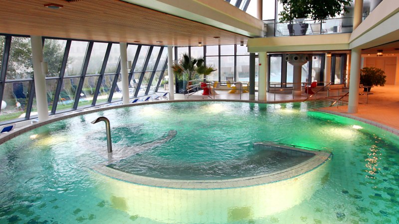 Modernes Schwimmbad mit großen Fenstern, grünem Wasser und Liegestühlen. Innenbereich mit Pflanzen und Sitzgelegenheiten., © Ufuk Arslan