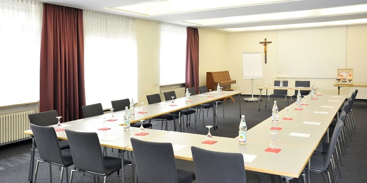 Konferenzraum mit U-förmiger Tischanordnung, Stühlen, Wasserflaschen und Gläsern. Ein Kreuz an der Wand, Flipchart und Klavier im Hintergrund., © Christkönigshaus Konferenzraum mit U-förmiger Tischanordnung, Stühlen, Wasserflaschen und Gläsern. Ein Kreuz an der Wand, Flipchart und Klavier im Hintergrund., © Christkönigshaus