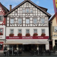 Fachwerkhaus mit Bäckerei und Café Beckabeck. Rote Blumen schmücken die Fenster. Vor dem Gebäude stehen Tische und Stühle unter Sonnenschirmen.
