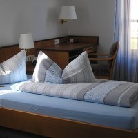 Ein Hotelzimmer mit Einzelbett, blauer Bettwäsche, Schreibtisch und Stuhl. Licht fällt durch gestreifte Vorhänge., © Hotel am Brunnen Ein Hotelzimmer mit Einzelbett, blauer Bettwäsche, Schreibtisch und Stuhl. Licht fällt durch gestreifte Vorhänge., © Hotel am Brunnen
