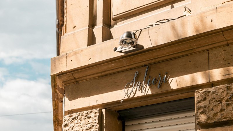 Fassade eines Geb&auml;udes mit dem Schriftzug 'Salieri' und einer &Uuml;berwachungskamera. Die Sonne wirft Schatten auf die sandfarbene Wand., &copy; SMG, Sarah Schmid