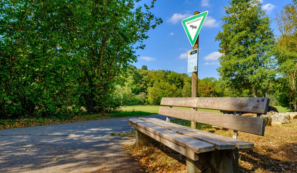 Holzbank am Wegesrand im Naturschutzgebiet, umgeben von grünen Bäumen und einem Hinweisschild. Der Himmel ist blau mit wenigen Wolken., © Remstal Tourismus e.V. Holzbank am Wegesrand im Naturschutzgebiet, umgeben von grünen Bäumen und einem Hinweisschild. Der Himmel ist blau mit wenigen Wolken., © Remstal Tourismus e.V.