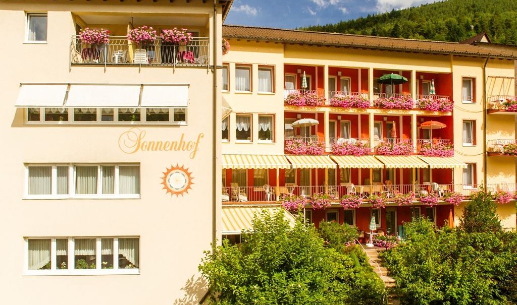 Das Hotel Sonnenhof zeigt eine Fassade mit bl&uuml;henden Balkonen und Sonnenschirmen. Der Schriftzug 'Sonnenhof' ist sichtbar. Im Hintergrund sind B&auml;ume zu sehen., &copy; Hotel Sonnenhof