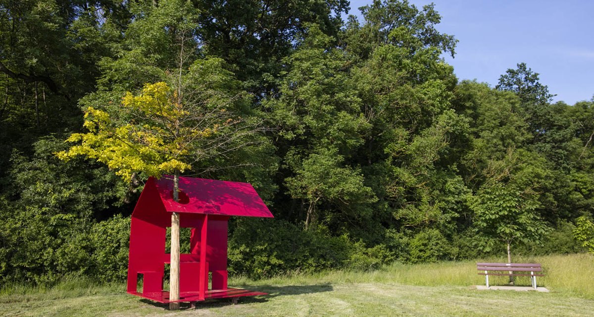 Eine rote Skulptur mit einem Baum in der Mitte steht auf einer Wiese vor einem dichten Wald. Daneben eine Bank und ein weiterer Baum., © Achim Mende Eine rote Skulptur mit einem Baum in der Mitte steht auf einer Wiese vor einem dichten Wald. Daneben eine Bank und ein weiterer Baum., © Achim Mende