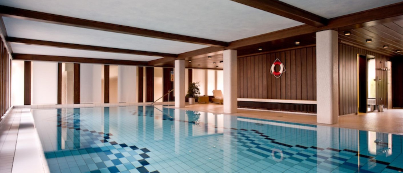 Ein leeres Hallenbad mit blauem Wasser, Holzwänden und einem Rettungsring an der Wand. Die Decke ist mit Holzbalken verziert., © Ringhotel Adler Asperg