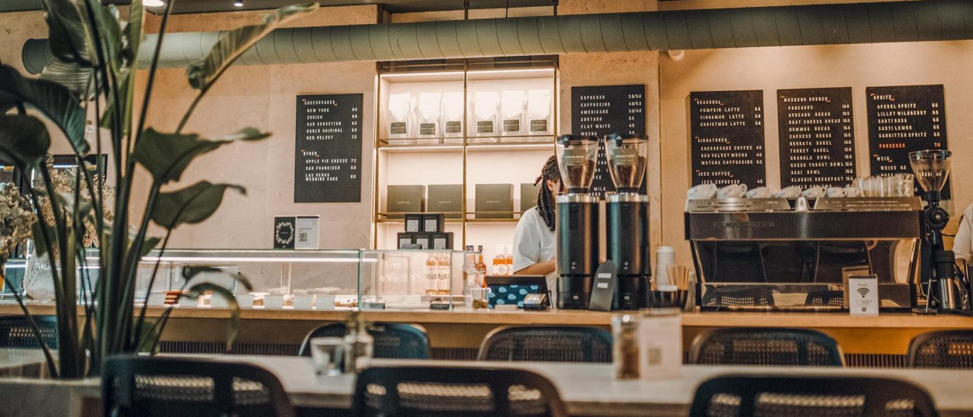 Modernes Café mit Theke, Kaffeemaschine und Speisekarten an der Wand. Pflanzen im Vordergrund, gemütliche Atmosphäre., © Maison Karl, Stuttgart Modernes Café mit Theke, Kaffeemaschine und Speisekarten an der Wand. Pflanzen im Vordergrund, gemütliche Atmosphäre., © Maison Karl, Stuttgart