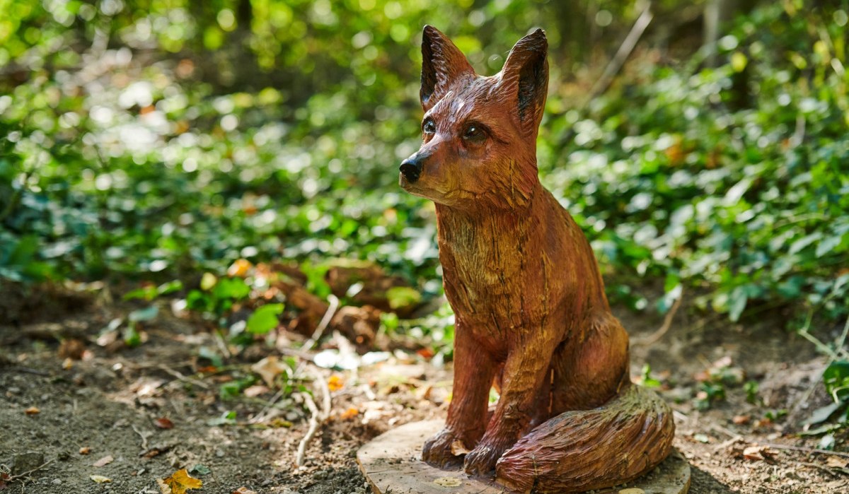 Eine detailreiche Holzskulptur eines sitzenden Fuchses im Wald, umgeben von gr&uuml;nem Laub und Sonnenlicht., &copy; Natur.Nah. Sch&ouml;nbuch & Heckeng&auml;u