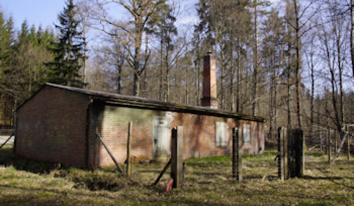 Ein altes Backsteingebäude mit Schornstein steht in einem lichten Wald. Ein verfallener Zaun umgibt das Gebäude. Ein altes Backsteingebäude mit Schornstein steht in einem lichten Wald. Ein verfallener Zaun umgibt das Gebäude.