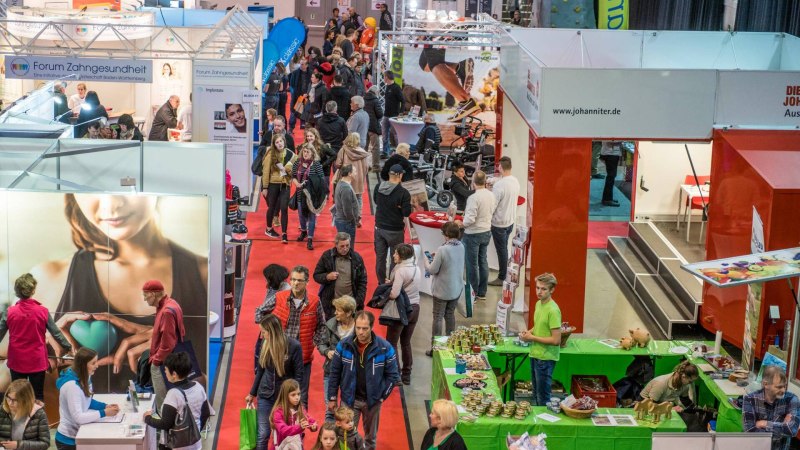 Menschen auf der vitawell Messe mit St&auml;nden zu Zahngesundheit und anderen Themen. Roter Teppich f&uuml;hrt durch die Halle.