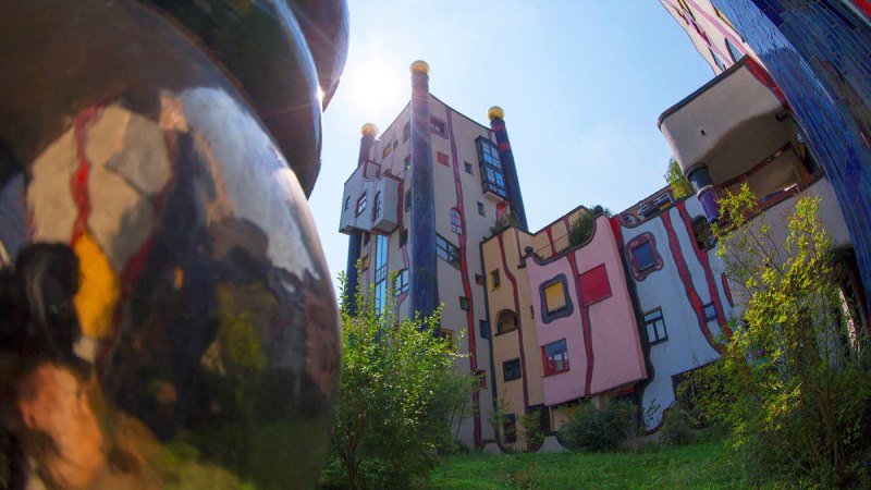 Bunte, unregelmäßige Gebäude mit runden Formen und goldenen Kuppeln im Hundertwasser-Stil, umgeben von Grünflächen., © Plochingen Bunte, unregelmäßige Gebäude mit runden Formen und goldenen Kuppeln im Hundertwasser-Stil, umgeben von Grünflächen., © Plochingen