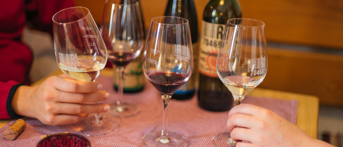 Zwei Personen halten Weingläser mit Rot- und Weißwein, stoßen an. Im Hintergrund stehen Weinflaschen auf einem Tisch mit lila Tischdecke., © 70469R! Weinbau Fabian Rajtschan