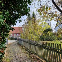 Ein gepflasterter Weg führt durch eine grüne Umgebung zu einem Fachwerkhaus und einem Kirchturm. Herbstliche Blätter und ein Holzzaun säumen den Weg., © Kulturamt Plochingen