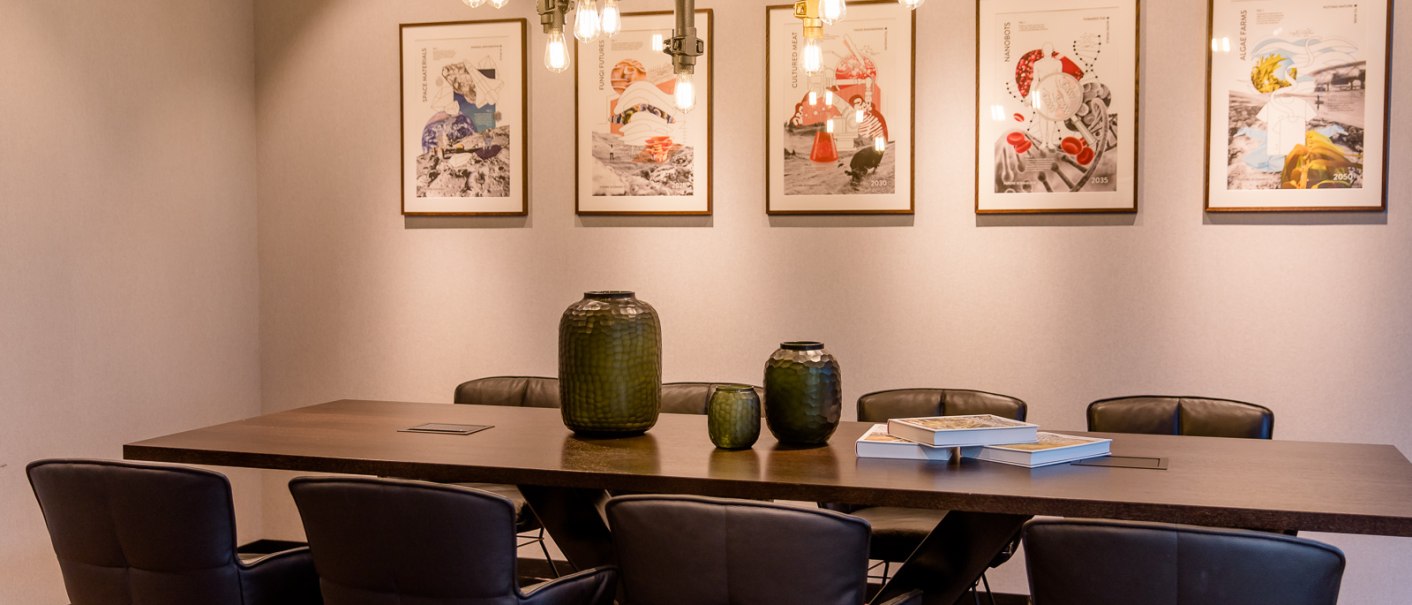Ein stilvoller Konferenzraum mit einem Holztisch, schwarzen Stühlen und modernen Hängelampen. An der Wand hängen fünf gerahmte Kunstwerke., © TOMAS