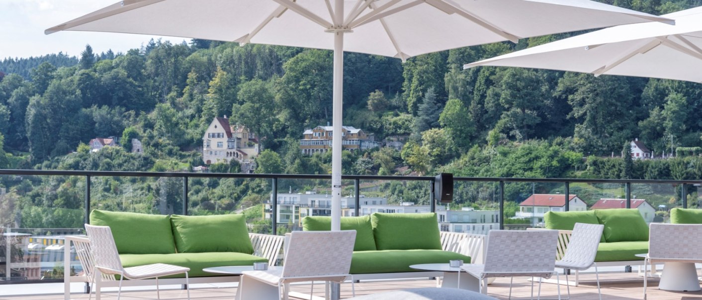 Rooftop-Bar mit grünen Sofas, weißen Tischen und Sonnenschirmen. Im Hintergrund bewaldete Hügel und Häuser. Entspannte Atmosphäre., © Hotel am Remspark Rooftop-Bar mit grünen Sofas, weißen Tischen und Sonnenschirmen. Im Hintergrund bewaldete Hügel und Häuser. Entspannte Atmosphäre., © Hotel am Remspark