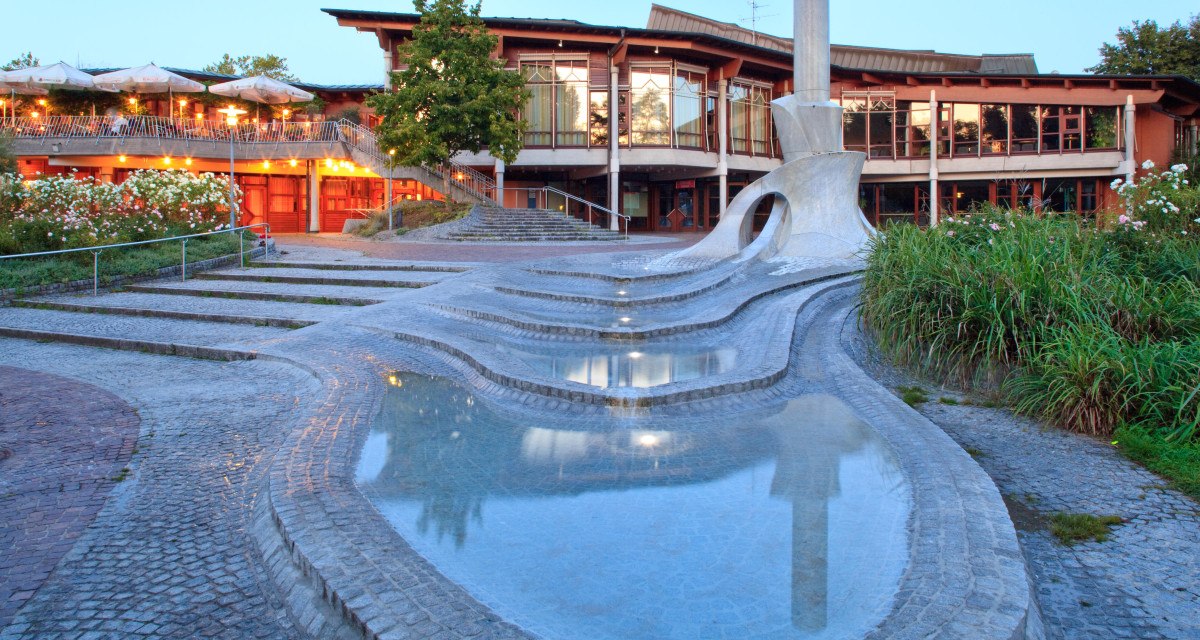 Moderner Brunnen mit geschwungenen Formen und Wasserbecken vor einem beleuchteten Gebäude mit Terrasse und Pflanzen., © Waiblingen - Stuttgart-Marketing GmbH Moderner Brunnen mit geschwungenen Formen und Wasserbecken vor einem beleuchteten Gebäude mit Terrasse und Pflanzen., © Waiblingen - Stuttgart-Marketing GmbH