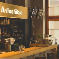 Gemütliche Bar mit Holztheke, Flaschenregal und Schild 'Bräustüble'. Schwarzes Brett kündigt Live-Musik an. Fenster im Hintergrund.