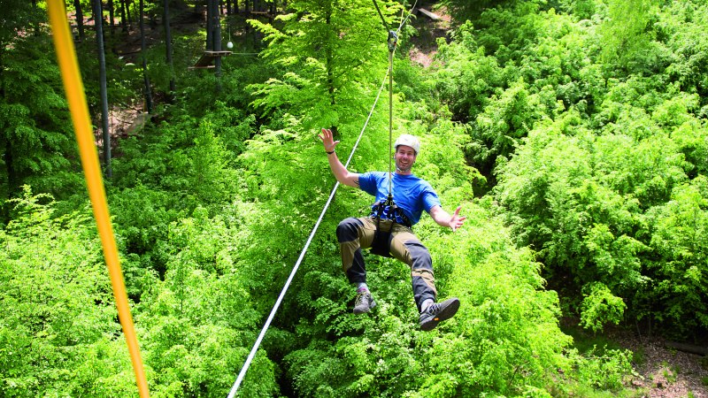 Person mit Helm und Kletterausr&uuml;stung schwebt an einem Seil &uuml;ber einem dichten, gr&uuml;nen Wald im Kletterwald Plochingen., &copy; Sebastian Berger