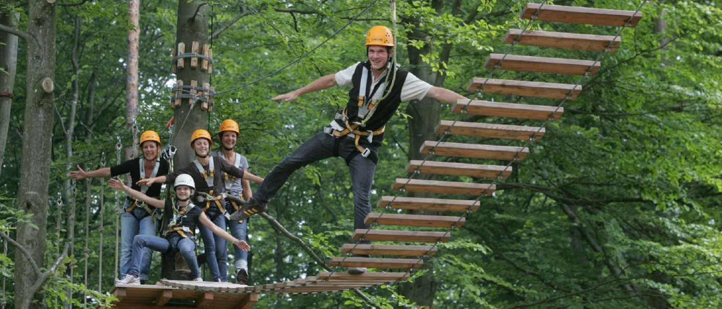 Menschen mit Helmen und Sicherheitsgurten auf einer Hängebrücke im Waldseilgarten Herrenberg, umgeben von grünen Bäumen., © Waldseilgarten Herrenberg