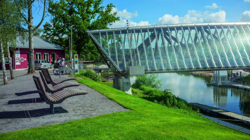 Moderne Glasbrücke in Remseck am Neckar, umgeben von grüner Landschaft und einem roten Gebäude. Sitzbänke und Fahrradständer im Vordergrund. Moderne Glasbrücke in Remseck am Neckar, umgeben von grüner Landschaft und einem roten Gebäude. Sitzbänke und Fahrradständer im Vordergrund.