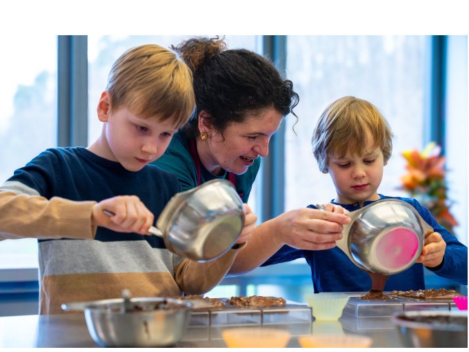 Zwei Kinder und eine Frau gie&szlig;en Schokolade in Formen. Sie sind in einer K&uuml;che mit gro&szlig;en Fenstern., &copy; Alfred Ritter GmbH &amp; Co. KG | Waldenbuch