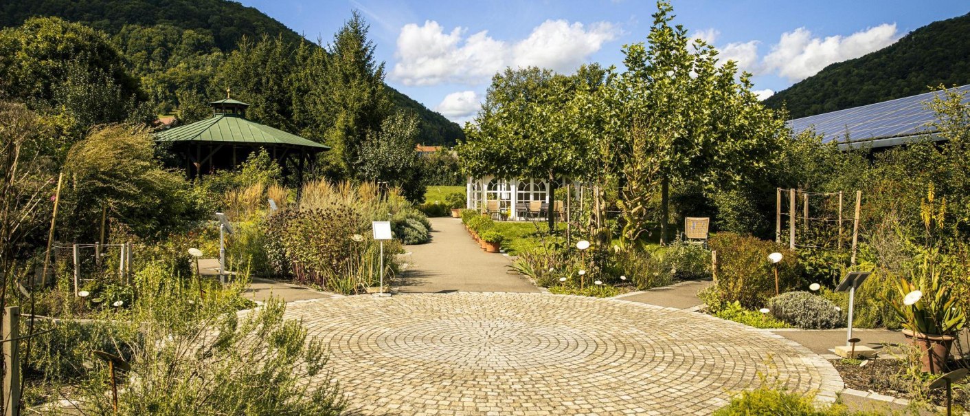 Kräutergarten mit gepflastertem Weg, üppiger Vegetation und Pavillon. Im Hintergrund bewaldete Hügel und blauer Himmel mit Wolken., © Stuttgart-Marketing GmbH, Sarah Schmid Kräutergarten mit gepflastertem Weg, üppiger Vegetation und Pavillon. Im Hintergrund bewaldete Hügel und blauer Himmel mit Wolken., © Stuttgart-Marketing GmbH, Sarah Schmid