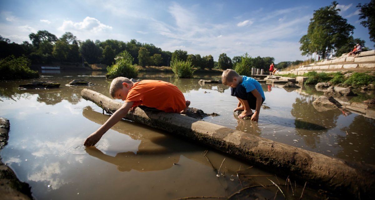 Zwei Kinder spielen am Ufer eines Flusses. Eines liegt auf einem Baumstamm, das andere kniet im Wasser. Im Hintergrund sind Bäume und ein sonniger Himmel., © Stoppel Zwei Kinder spielen am Ufer eines Flusses. Eines liegt auf einem Baumstamm, das andere kniet im Wasser. Im Hintergrund sind Bäume und ein sonniger Himmel., © Stoppel