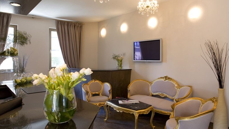 Elegante Lobby mit goldenen Möbeln, einem Tisch, Blumenarrangements und einem Fernseher an der Wand. Dezente Beleuchtung und Vorhänge., © Hotel das kleine Ritz Elegante Lobby mit goldenen Möbeln, einem Tisch, Blumenarrangements und einem Fernseher an der Wand. Dezente Beleuchtung und Vorhänge., © Hotel das kleine Ritz
