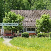 Biergarten Maisentalstüble mit roten Tischen und Stühlen, umgeben von grüner Natur und blühenden Wiesen. Biergarten Maisentalstüble mit roten Tischen und Stühlen, umgeben von grüner Natur und blühenden Wiesen.