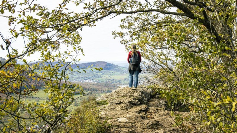 Eine Person steht auf dem Beurener Fels, umgeben von Bäumen, und blickt auf die weite Landschaft von Beuren., © SMG, Sarah Schmid Eine Person steht auf dem Beurener Fels, umgeben von Bäumen, und blickt auf die weite Landschaft von Beuren., © SMG, Sarah Schmid