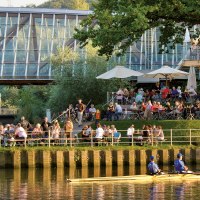 Menschen genießen das Wetter in einem Café am Flussufer, während ein Ruderboot vorbeifährt. Im Hintergrund ist eine moderne Brücke zu sehen., © Michael Fuchs