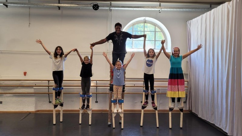 Kinder und ein Erwachsener stehen auf Stelzen in einem Raum. Sie halten sich an den Händen und lächeln. Im Hintergrund sind Vorhänge und Fenster zu sehen., © Kunstschule Labyrinth Kinder und ein Erwachsener stehen auf Stelzen in einem Raum. Sie halten sich an den Händen und lächeln. Im Hintergrund sind Vorhänge und Fenster zu sehen., © Kunstschule Labyrinth